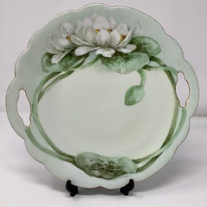 May include: A round, scalloped-edge serving plate with a pale green background. The plate features a hand-painted design of white water lilies with yellow centers and green leaves. The plate has two handle cutouts.