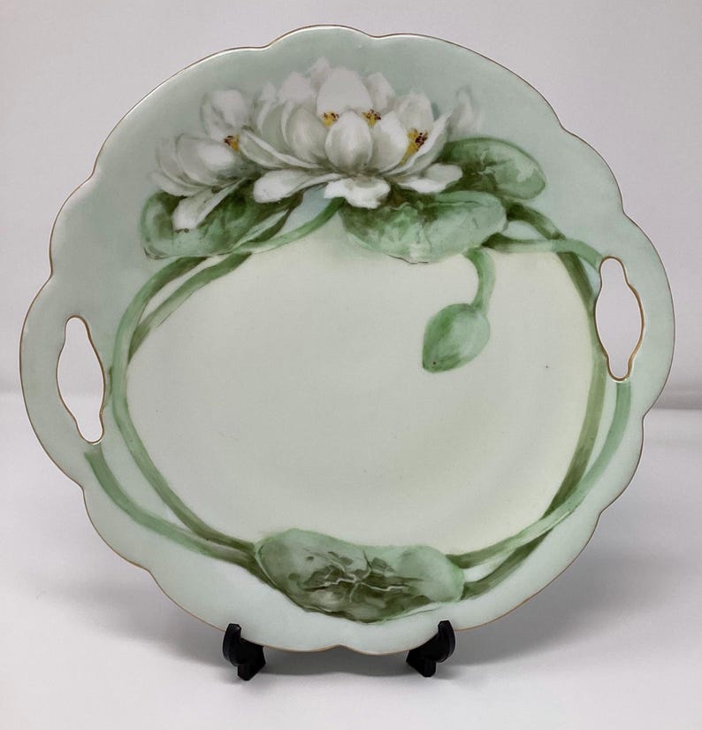 May include: A round, scalloped-edge serving plate with a pale green background. The plate features a hand-painted design of white water lilies with yellow centers and green leaves. The plate has two handle cutouts.