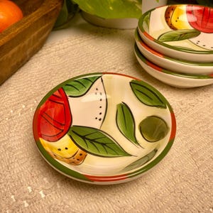 Puede incluir: Un juego de cuatro cuencos peque&ntilde;os de cer&aacute;mica pintados a mano con un dise&ntilde;o de frutas y hojas. Los cuencos tienen una base blanca con detalles en rojo, amarillo y verde. Los bordes son verdes y rojos.