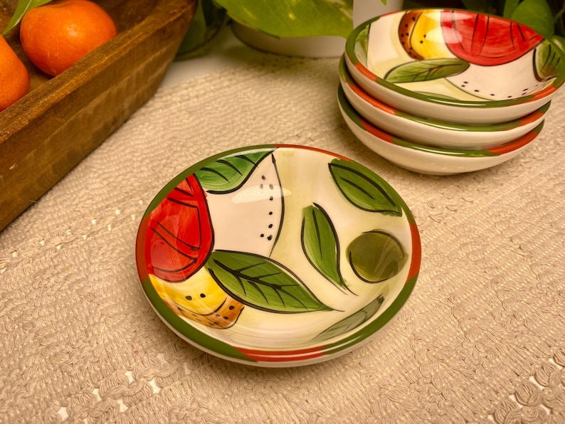 Puede incluir: Un juego de cuatro cuencos peque&ntilde;os de cer&aacute;mica pintados a mano con un dise&ntilde;o de frutas y hojas. Los cuencos tienen una base blanca con detalles en rojo, amarillo y verde. Los bordes son verdes y rojos.