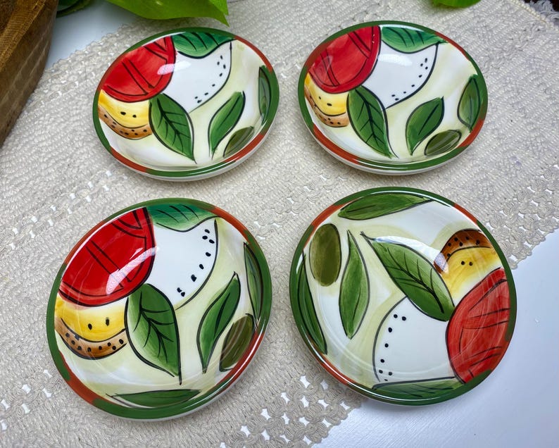 Puede incluir: Cuatro peque&ntilde;os cuencos de cer&aacute;mica pintados a mano. Cada cuenco presenta un interior blanco con un dise&ntilde;o de elementos rojos, amarillos y verdes, incluyendo hojas y frutas. Los bordes son verdes y los cuencos est&aacute;n dispuestos sobre una superficie de tono neutro.