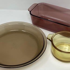 May include: A collection of vintage glass bakeware in shades of brown and amber. The set includes a rectangular loaf pan, a round pie dish, and a small round casserole dish, all with handles. The pieces are arranged on a white surface.