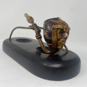 May include: A vintage tobacco pipe with a carved Native American head bowl. The pipe is supported by a metal stand and rests on a black wooden base. The pipe's stem and stand have a brass finish.