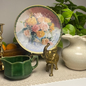 May include: A still life arrangement featuring a decorative plate with a floral design, a brass candlestick, a green creamer, a small brass elephant figurine, and a speckled white pitcher. Oranges are in a wooden box.