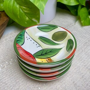 Puede incluir: Una pila de cuatro platos peque&ntilde;os y redondos de cer&aacute;mica. Cada plato presenta un dise&ntilde;o pintado de hojas verdes, detalles rojos y amarillos y un borde verde. Los platos est&aacute;n apilados sobre una superficie blanca texturizada.