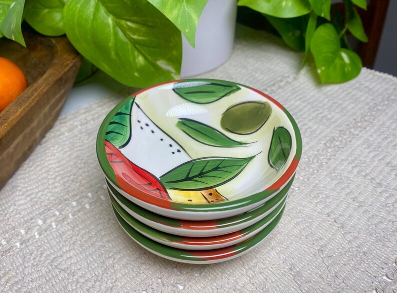 Puede incluir: Una pila de cuatro platos peque&ntilde;os y redondos de cer&aacute;mica. Cada plato presenta un dise&ntilde;o pintado de hojas verdes, detalles rojos y amarillos y un borde verde. Los platos est&aacute;n apilados sobre una superficie blanca texturizada.