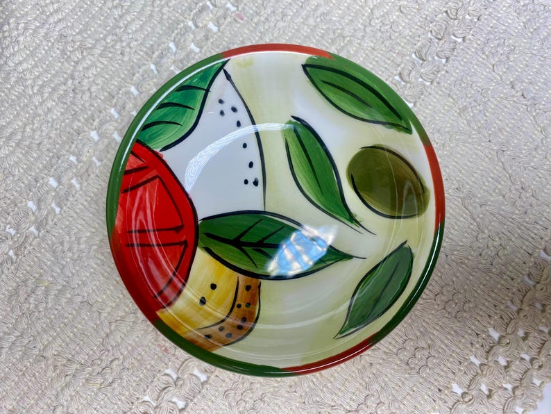 Puede incluir: Un cuenco de cer&aacute;mica pintado a mano con un colorido dise&ntilde;o de frutas y hojas. El cuenco presenta una paleta de colores rojo, verde, amarillo y blanco. El borde es verde con un acento rojo. El cuenco es adecuado para servir alimentos.