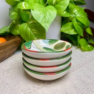 Puede incluir: Una pila de cuatro cuencos peque&ntilde;os de cer&aacute;mica blanca. Cada cuenco tiene un borde verde y rojo, con un dise&ntilde;o pintado de hojas verdes y detalles amarillos en el interior. Los cuencos est&aacute;n apilados sobre una superficie blanca texturizada.