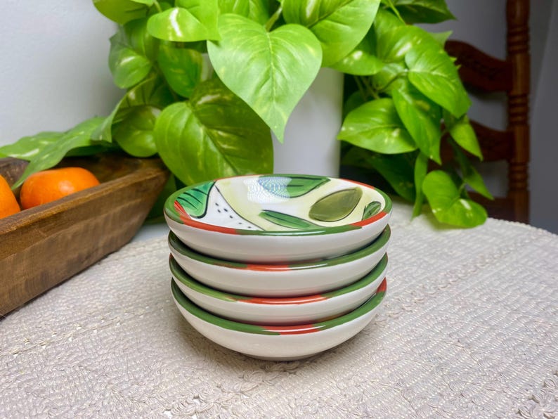 Puede incluir: Una pila de cuatro cuencos peque&ntilde;os de cer&aacute;mica blanca. Cada cuenco tiene un borde verde y rojo, con un dise&ntilde;o pintado de hojas verdes y detalles amarillos en el interior. Los cuencos est&aacute;n apilados sobre una superficie blanca texturizada.