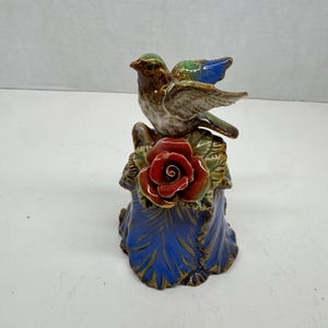 May include: A ceramic bird figurine with a brown body, green head, and blue wings perched atop a red rose. The base is blue with gold accents. The figurine is a decorative item.