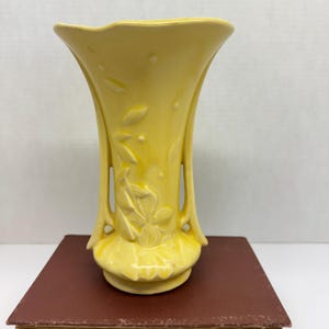May include: A yellow ceramic vase with a flared top and a decorative floral design. The vase has a pedestal base and two handles. The vase is approximately 8 inches tall.