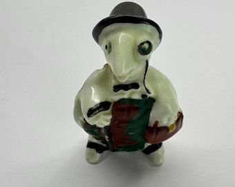 May include: A small, ceramic figurine of a creature wearing a black hat, bow tie, and apron. The figurine has a light green body, black and white striped legs, and is holding a basket. The apron is green and red.