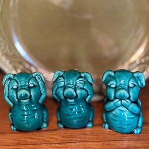 May include: Three teal ceramic figurines of seated pigs, each with a different gesture: covering eyes, ears, and mouth. The figurines are glossy and have detailed facial features. They are arranged on a wooden surface.