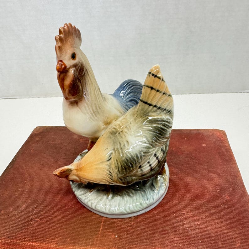 May include: A ceramic figurine of a rooster and hen. The rooster has a red comb and wattle, with blue and white feathers. The hen has brown, tan, and black feathers. The figurine is on a white base with green grass detailing.