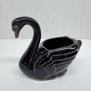 May include: A glossy black ceramic swan planter featuring a long, curved neck and a hollow body with molded feather details. The swan has a small red accent on its beak and a yellow eye detail, with a smooth, reflective finish throughout the piece.