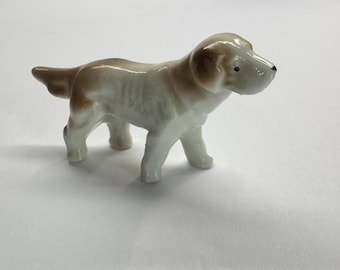 May include: A ceramic figurine of a dog in a standing pose. The dog has a white body with brown markings on its head, ears, and tail. The figurine has a glossy finish and is set against a white background.
