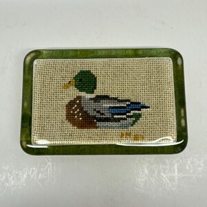 May include: A rectangular, green-framed glass object featuring a cross-stitch design of a mallard duck. The duck has a green head, brown body, and blue wings. The background is a tan, textured fabric.