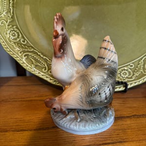 May include: A ceramic rooster and hen figurine. The rooster has a red comb and wattle, with a white and tan body. The hen is gray and brown with black markings. The base is a light blue color.