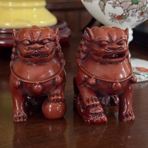 May include: Two red-brown Foo Dog statues. Each statue is in a seated position with a fierce expression, detailed fur, and a ball under one paw. The statues are on a dark wooden surface.