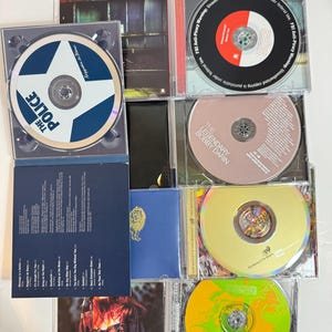 May include: A collection of eight compact discs (CDs) in jewel cases. The CDs feature various artists, including The Police, Bobby Darin, and a band called "The Shadows". The CDs are arranged in two rows of four, with the cases open to reveal the discs.