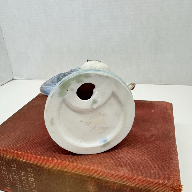May include: A ceramic figurine base showing a circular white underside with a central hole and a light blue glazed edge. The object is positioned on a red book cover. The base features a smooth, matte white finish with subtle blue and brown accents.