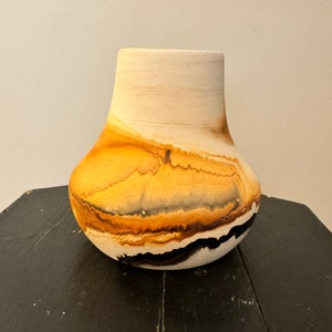 May include: A white ceramic vase with a rounded body and a wide mouth. The vase has a marbled design with shades of yellow, orange, brown, and black.