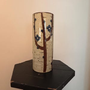 May include: A cylindrical ceramic vase with a beige speckled background. The vase features a brown tree branch design with blue berry clusters. The vase is displayed on a black, hexagonal table.