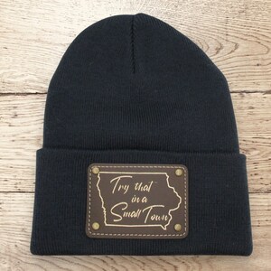 May include: A black knit beanie with a brown leather patch. The patch features the outline of Iowa and the text "Try that in a Small Town" in a gold color. The beanie is made of a soft, knitted material.