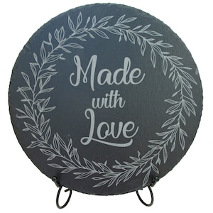 May include: A round black slate serving board with a white engraved wreath and the text "Made with Love". The board is on a black metal stand.