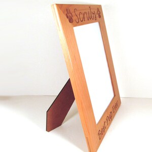 May include: A wooden picture frame with a light brown finish. The frame has a cutout of a paw print on each side and the text "Scruffy" at the top. The bottom of the frame has the text "Best Pup Ever".