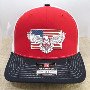 May include: Red, white, and blue trucker hat with an American flag and eagle design. The hat is adjustable and has a black mesh back. The hat has the text "Richardson 112 Original Trucker Adjustable" on the back.