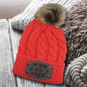 May include: Red knitted beanie with a faux fur pom pom and a brown leather patch that says "No Hair Don't Care".