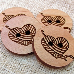 May include: Four wooden buttons with a heart-shaped design featuring a ball of yarn and knitting needles. The buttons are light brown and have a smooth finish.