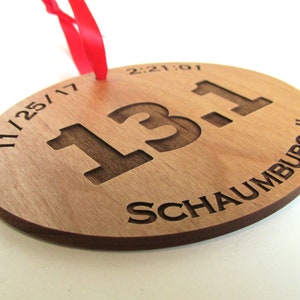 May include: A wooden round ornament with the text "11/25/17 13.1 Schaumburg, IL" and the time "2:21:01" engraved on it. The ornament is attached to a red ribbon.