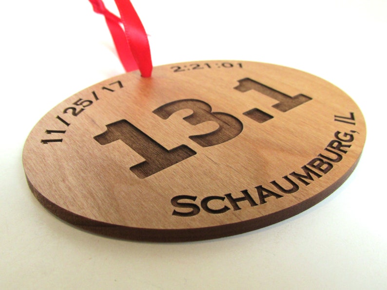 May include: A wooden round ornament with the text "11/25/17 13.1 Schaumburg, IL" and the time "2:21:01" engraved on it. The ornament is attached to a red ribbon.