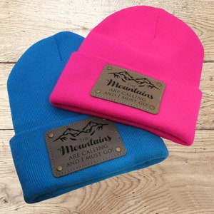 May include: Two knit beanies with leather patches. The patches are brown and feature a mountain range with the text "The Mountains Are Calling And I Must Go!" in black.