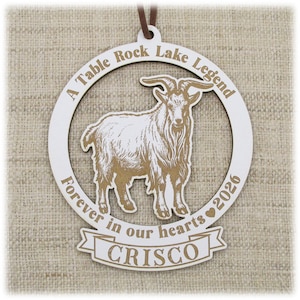 May include: A circular white wooden ornament featuring a brown engraved goat in the center. The text reads A Table Rock Lake Legend, Forever in our hearts 2026, and the name CRISCO on a banner at the bottom. A brown cord is attached at the top.