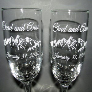 May include: Two clear glass champagne flutes with a mountain range design and the text "Chad and Anne January 31, 2020" etched on each.