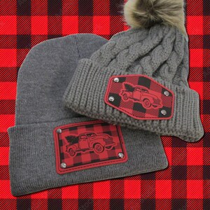 May include: Two gray knit winter hats with a red and black buffalo plaid design and a red truck with a Christmas tree. The truck is on a patch of faux leather with silver rivets.