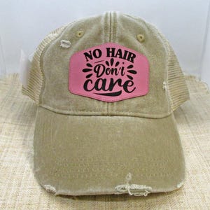 May include: A tan distressed trucker hat with a pink patch that says "No Hair Don't Care" in black lettering.