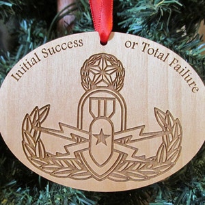 May include: A wooden Christmas ornament with a red ribbon. The ornament is shaped like an oval and features a stylized image of a bomb with wings and a star. The text "Initial Success or Total Failure" is engraved around the edge of the ornament.