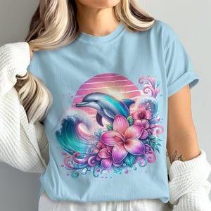 Pode incluir: Uma camiseta azul claro com uma estampa gr&aacute;fica de um golfinho saltando sobre uma onda com flores rosa e roxas. O gr&aacute;fico est&aacute; sobre um fundo de p&ocirc;r do sol.