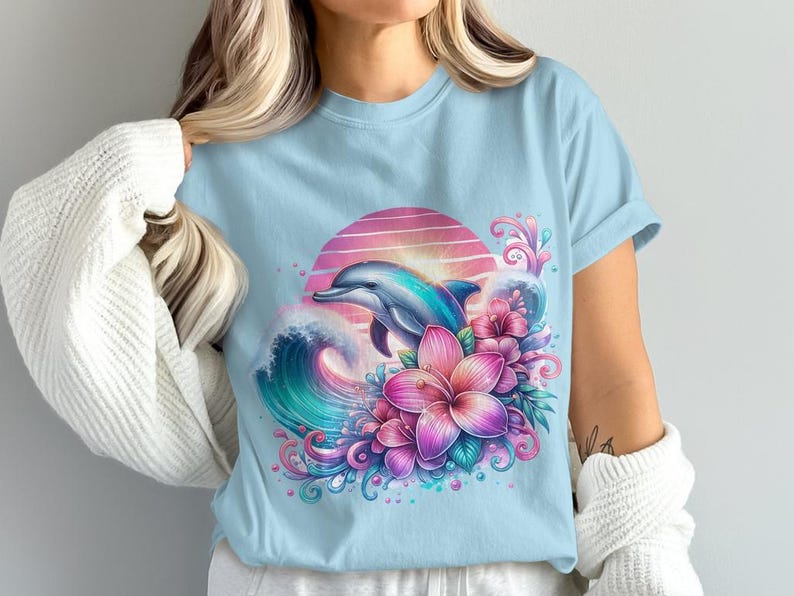 Pode incluir: Uma camiseta azul claro com uma estampa gr&aacute;fica de um golfinho saltando sobre uma onda com flores rosa e roxas. O gr&aacute;fico est&aacute; sobre um fundo de p&ocirc;r do sol.