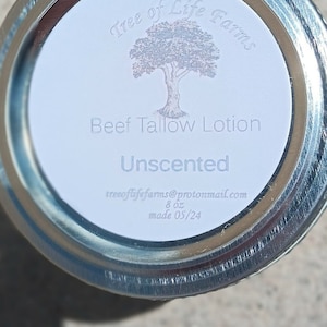 May include: A white label with the text "Tree of Life Farms" and a drawing of a tree. The label also says "Beef Tallow Lotion Unscented" and "8 oz made 05/24". The label is on a silver jar lid.