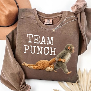 Puede incluir: Sudadera marrón con el texto "TEAM PUNCH" en blanco. La sudadera presenta un gráfico de un mono tirando de un peluche. Un sombrero marrón es visible en la esquina superior izquierda.
