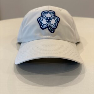 May include: A white baseball cap with a blue and white clover design on the front.