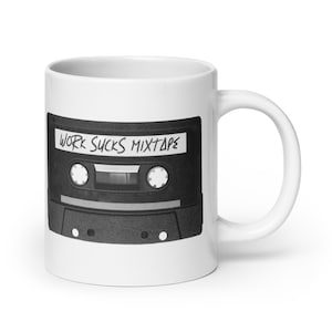 Könnte beinhalten: Weiße Keramik-Tasse mit einem schwarz-weißen Kassettenband-Design. Die Kassettenband-Grafik enthält den Text "WORK SUCKS MIXTAPE" in einer handschriftlichen Schriftart. Die Tasse hat einen Standardgriff.