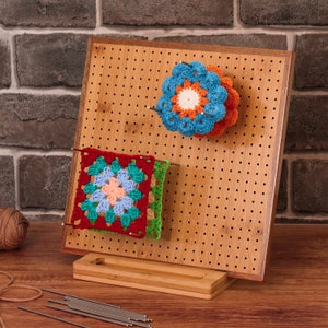 May include: A bamboo pegboard with a stand, used for displaying crocheted items. The pegboard has a variety of crocheted squares and flowers in red, green, blue, orange, and white.