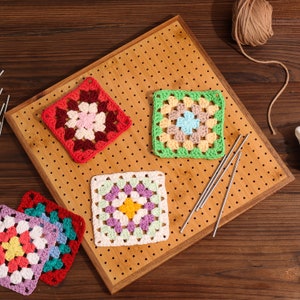 May include: Four colourful crochet granny squares on a wooden surface with knitting needles. The squares are in shades of red, green, white, and purple.