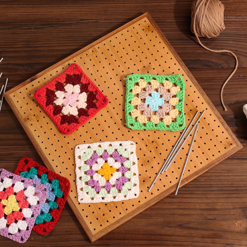 May include: Four colourful crochet granny squares on a wooden surface with knitting needles. The squares are in shades of red, green, white, and purple.